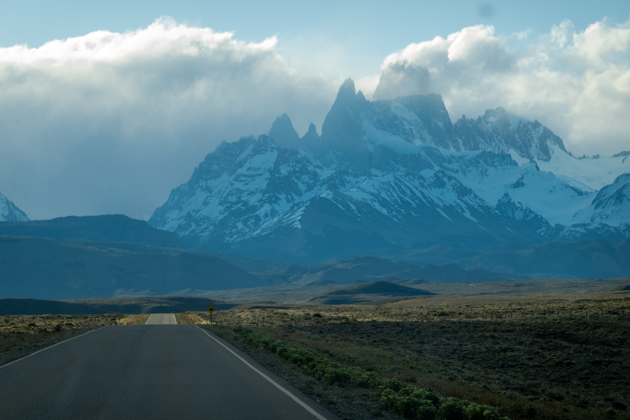 a road on the lower left heads towards some jagged mountain summits with snow
