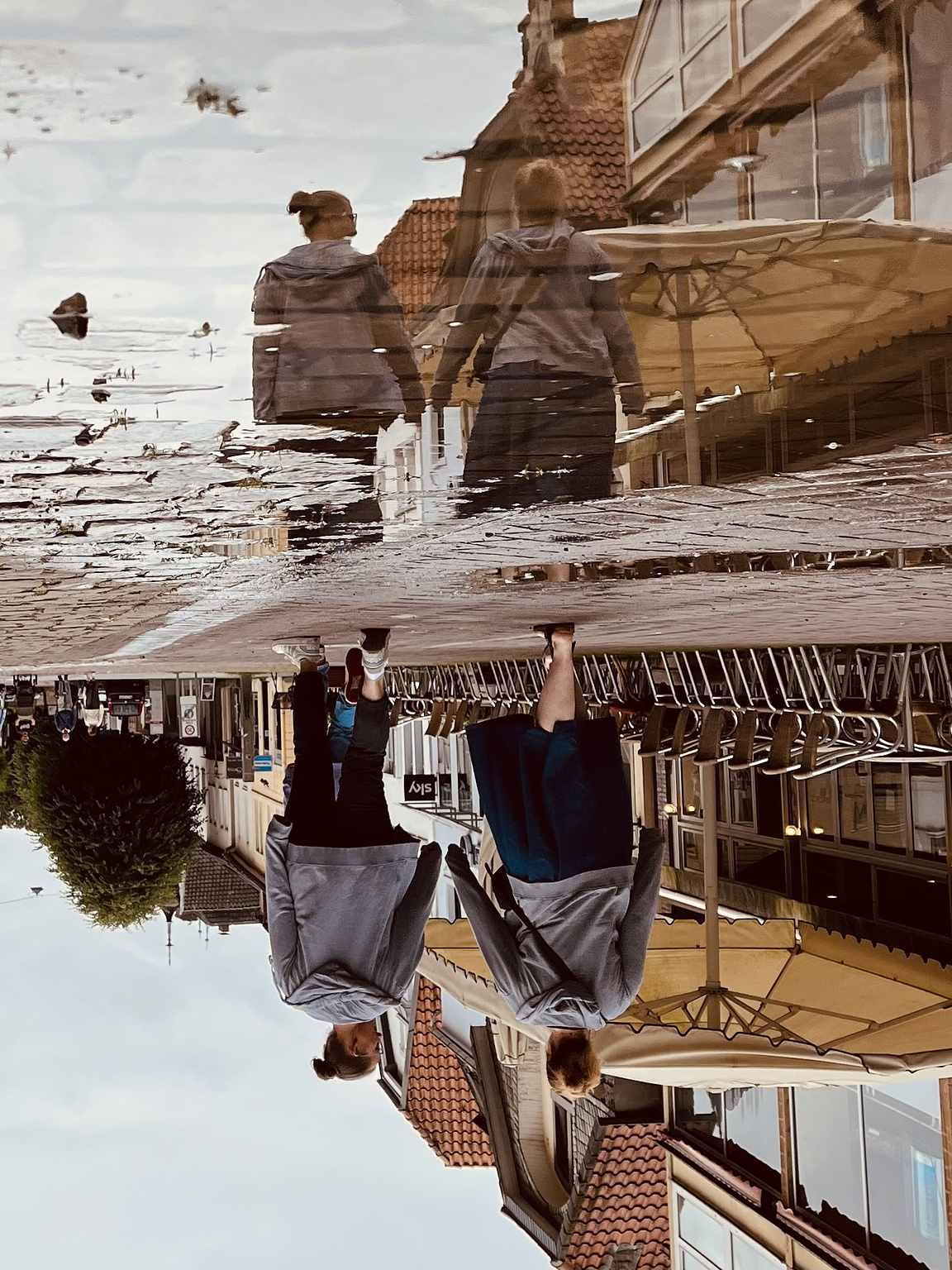 A couple of two women walking on a cobble stone street, reflected in a puddle on the street. on the right side of the picture there are some chairs and tishes in front of a cafe, and yellow sun shades. The whole picture is turned upside down.