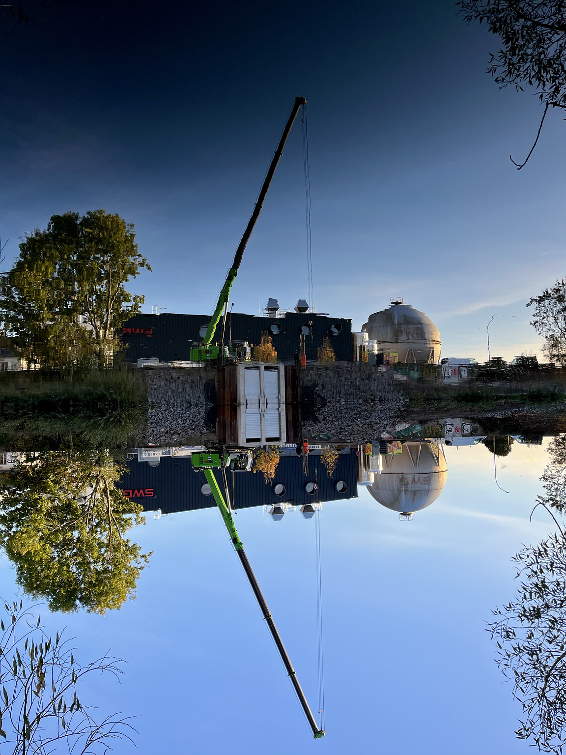 Construction crane and trees, reflected in the surface of a river. Gentlemans evening light (Blue Hour). The whole picture is turned upsidedown.