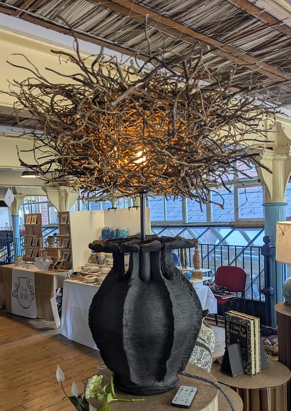 A lamp base made of black clay, in the stylised form of a poppy seed head, topped with a fantastic looking, and very large, 'birds nest' lamp shade (makers link in above text)
