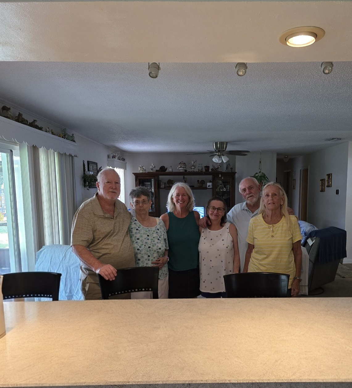 A group of six older adults, smiling and standing together in a warmly lit living room. They’re posing in front of a kitchen counter with chairs, and family photos and decorations are visible in the background. The photographer is not pictured.