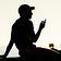 A guy in silhouette against a slightly warm toned sky, sat on a beach wall with one hand behind propping him up, and the other holding a phone in front of him. There is a glass of cider balanced on the wall top in front of him, which the light is shining through, so appears as the only spot of amber colour.