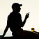 A guy in silhouette against a slightly warm toned sky, sat on a beach wall with one hand behind propping him up, and the other holding a phone in front of him. There is a glass of cider balanced on the wall top in front of him, which the light is shining through, so appears as the only spot of amber colour.