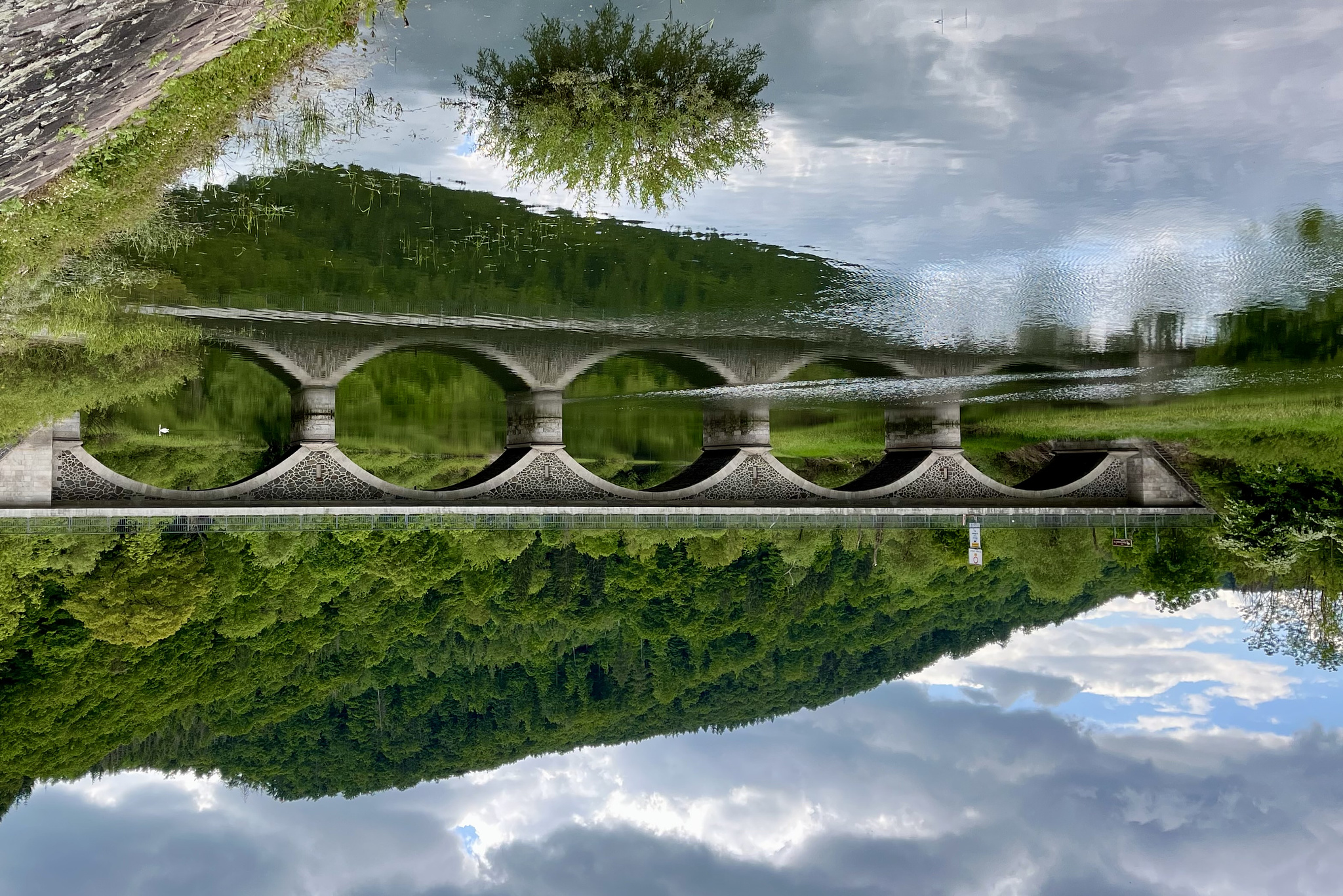 An older bridge with five arches, reflected in the water. Above a blue sky with some clouds. Behind the bridge there is a hill covered with green trees. On the left side there Are some green bushes. The whole picture is turned upside down.
