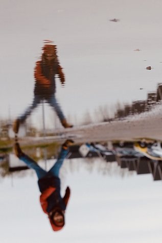 Reflection of a child wearing an orange-blue jacket jumping across a puddle on the street. The picture is turned upside down.