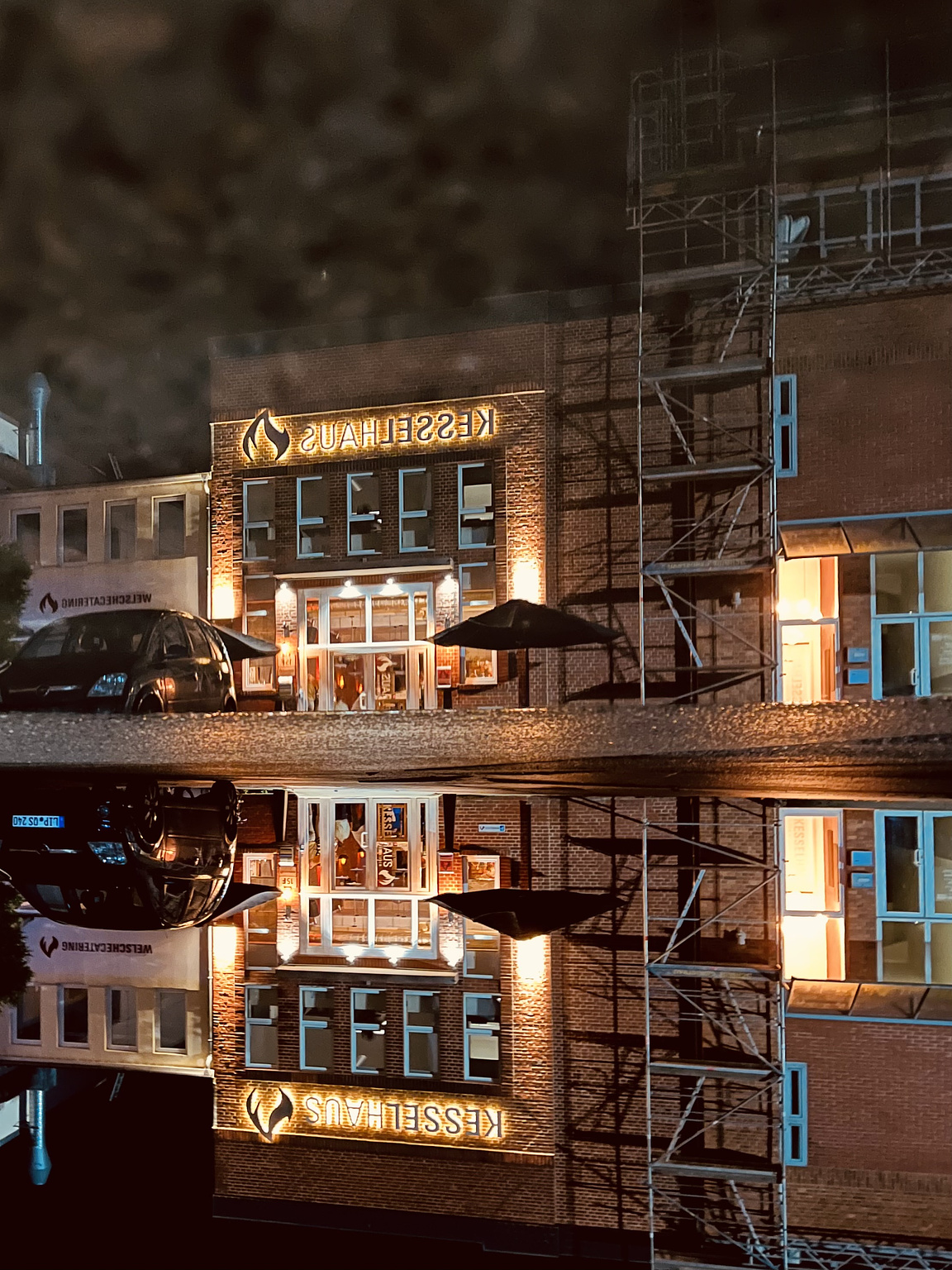 Old industry buildings, now used for events. It is night and there are some lights. The situatiom is reflected in a puddle. The whole picture is turned upsidedown.