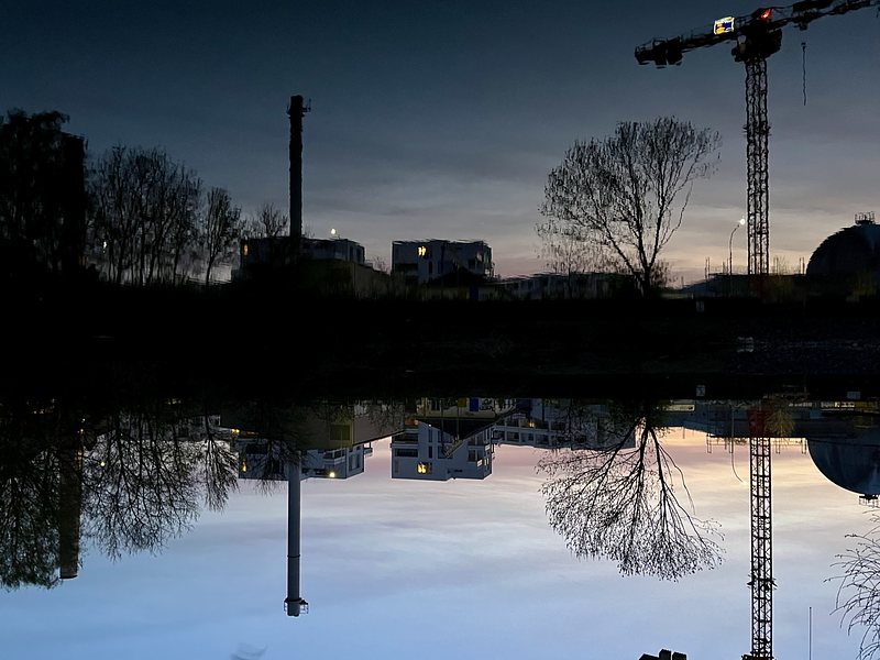 Public utility buildings, a few trees and a crane are reflected in a river. It's already dark, but there's a bit of daylight and evening glow in the sky. The picture is turned upside down.