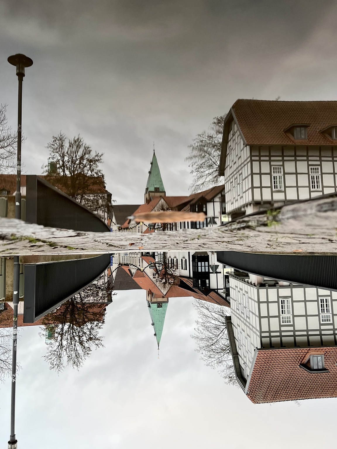 Cobblestone street with a church tower in the middle. In the foreground on the right is a half-timbered house. The scene is reflected in a puddle on the street. The entire image is upside down.
