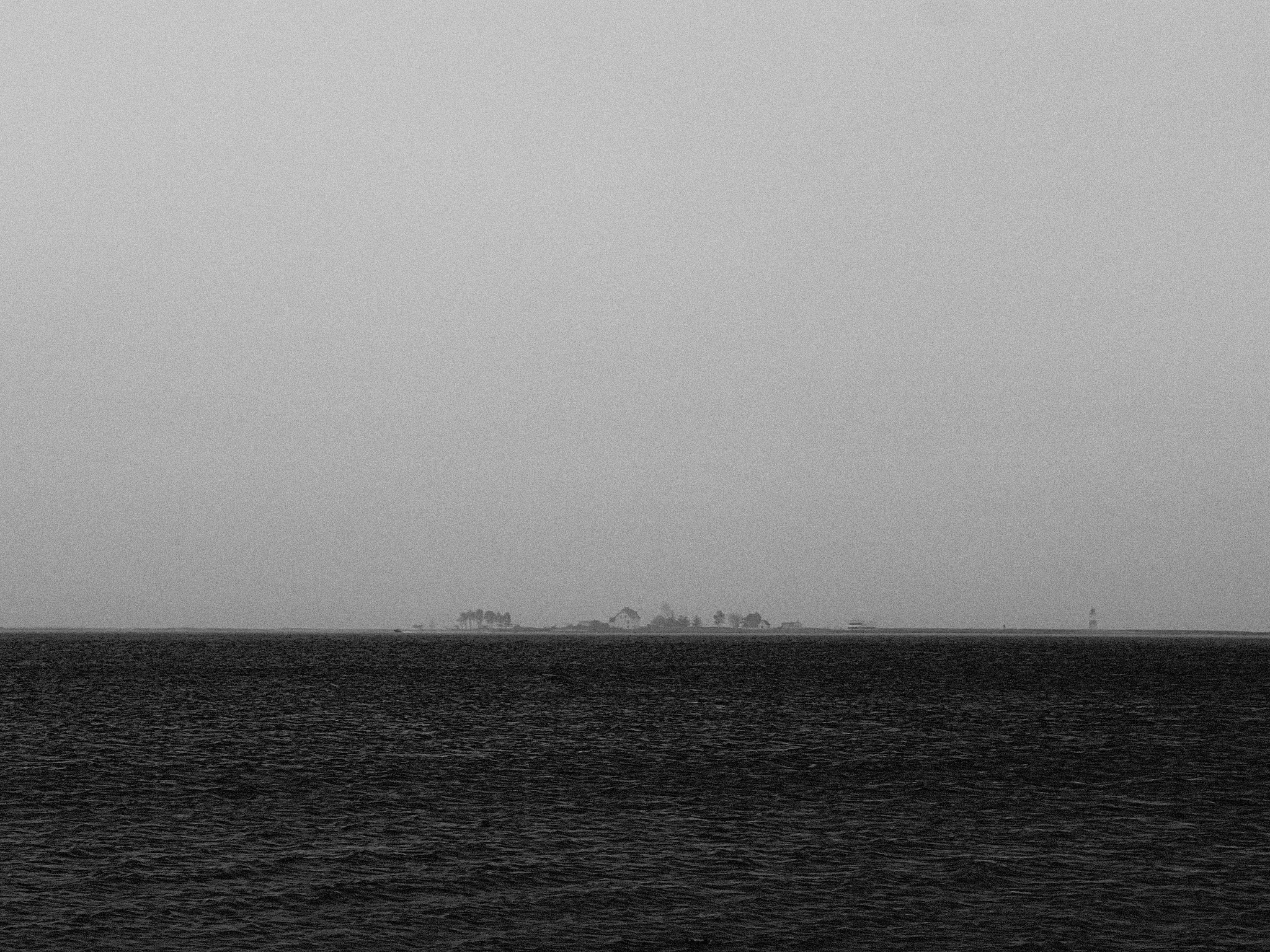 A black and white photograph of a distant shoreline, featuring a small, faint island with trees barely visible on the horizon. The foreground is dominated by a vast, dark sea with gentle ripples. The sky is overcast and merges smoothly with the water, creating a minimalist and atmospheric scene.