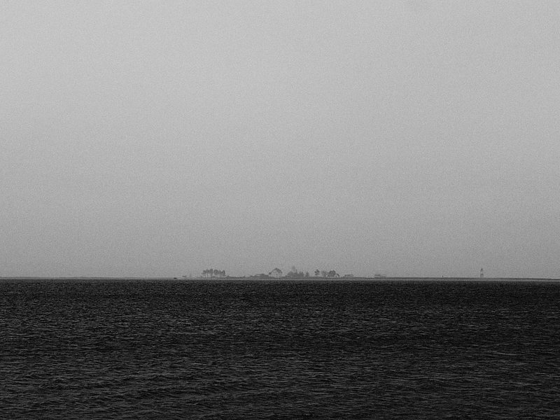 A black and white photograph of a distant shoreline, featuring a small, faint island with trees barely visible on the horizon. The foreground is dominated by a vast, dark sea with gentle ripples. The sky is overcast and merges smoothly with the water, creating a minimalist and atmospheric scene.