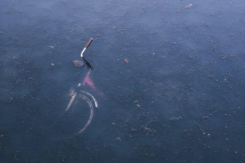 A bicycle is partially submerged in a body of water. Only the handlebars and part of the front wheel are visible above the surface, which is covered with patches of debris and reflections of the sky.