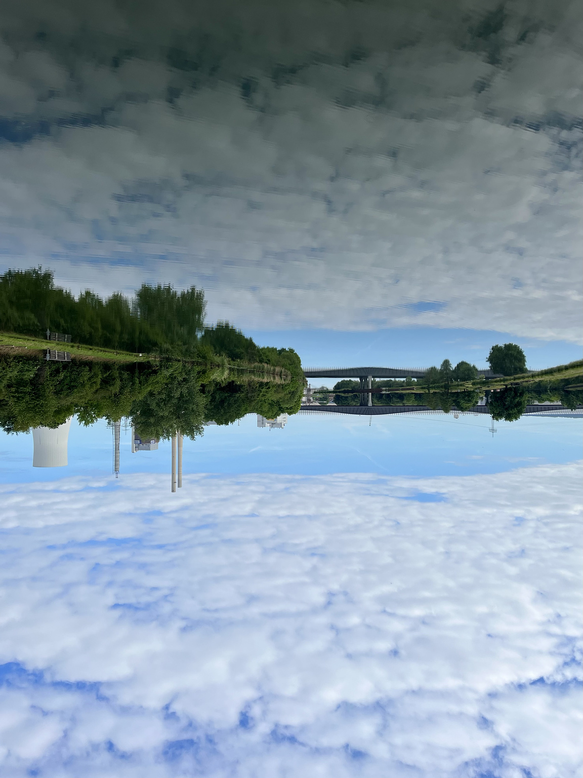 Cool power plant, a Bridge and some more industry buildings, also some trees, all reflected in a canal. Above it a Bit of Blue sky shining through a lot of white clouds. The Picture is turned upsidedown.