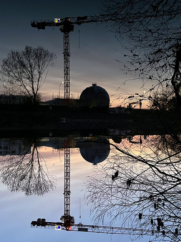 Public utility buildings, a few trees and a crane are reflected in a river. It's already dark, but there's a bit of daylight and evening glow in the sky. The picture is turned upside down.