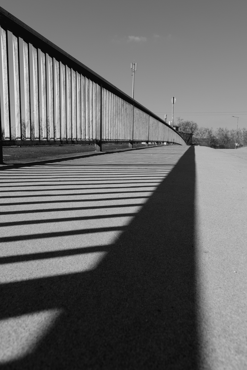 Black and white photo of a bridge with metal railings on the left side casting elongated shadows diagonally across a concrete walkway. The sky is clear, and there are trees and a few utility poles visible in the background.