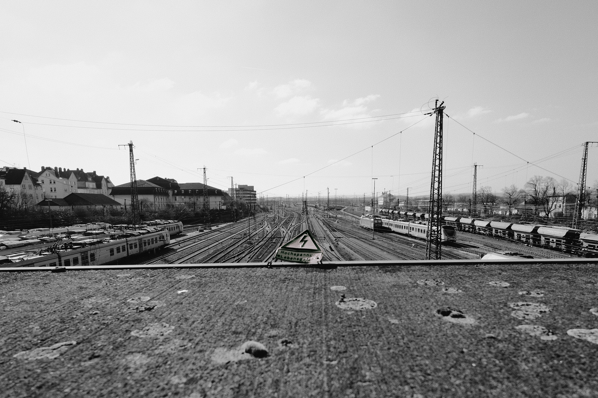 Black and white image of a railway yard with multiple train tracks extending into the distance. On the left, there are trains parked on some tracks, with a mix of freight and passenger cars visible on the right. Overhead power lines are strung between tall pylons. In the foreground, there is a concrete surface with some visible textures and a triangular warning sign featuring a lightning bolt symbol. Buildings and trees line the background under a clear sky.