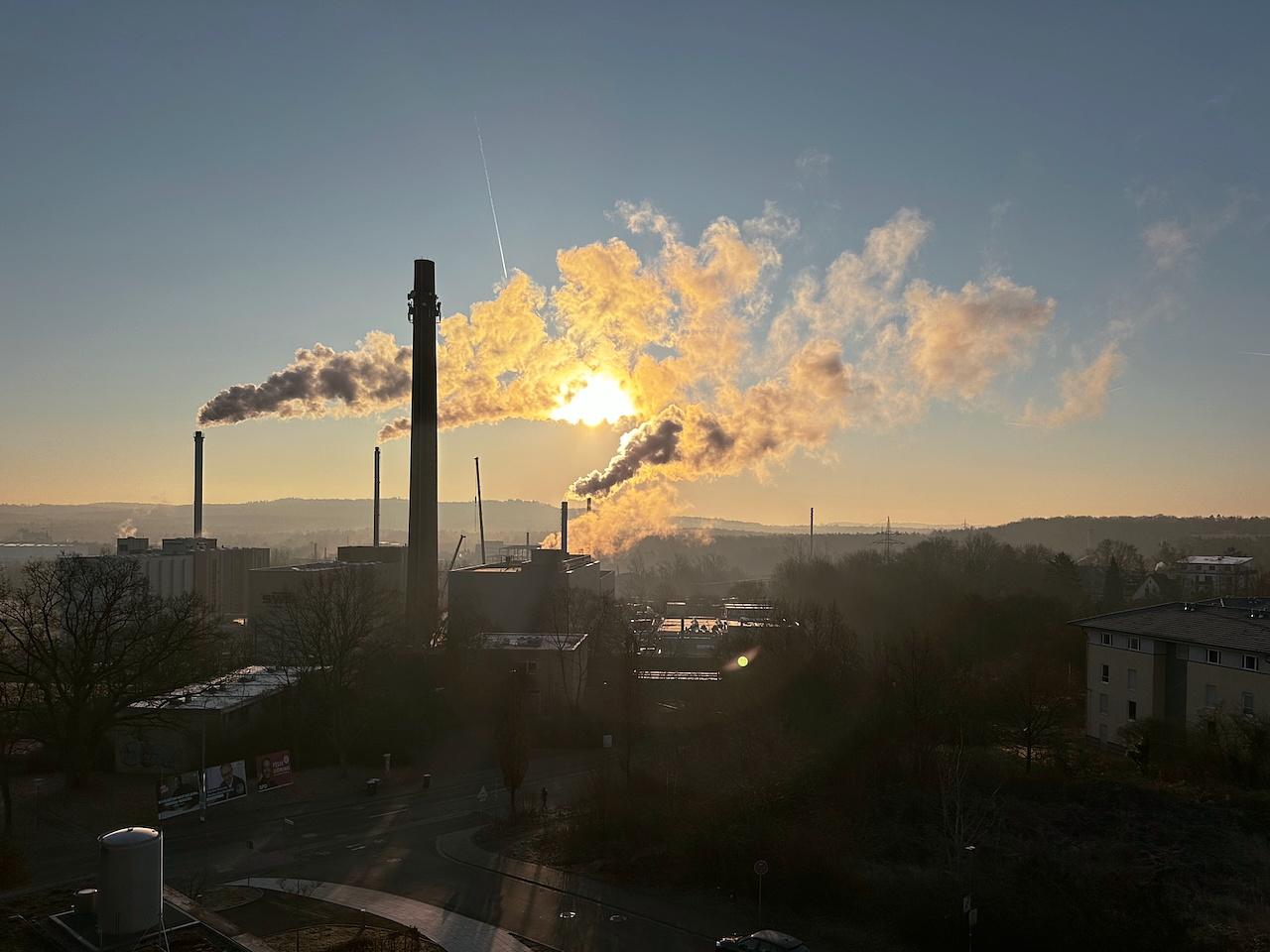 A factory with several tall smokestacks releasing smoke under a clear sky during sunrise or sunset. The sun is partially obscured by smoke, creating a warm glow. Surrounding the industrial complex are trees, buildings, and a road in the foreground. The landscape in the background is slightly hilly, with a layer of haze or mist.