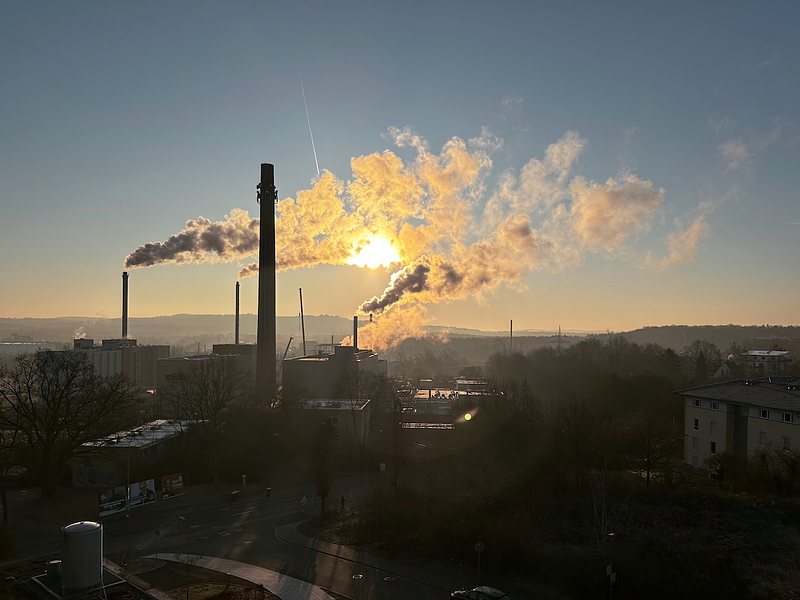 A factory with several tall smokestacks releasing smoke under a clear sky during sunrise or sunset. The sun is partially obscured by smoke, creating a warm glow. Surrounding the industrial complex are trees, buildings, and a road in the foreground. The landscape in the background is slightly hilly, with a layer of haze or mist.