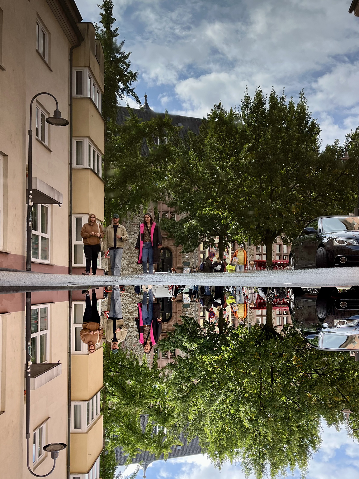 Street with yellow three storey buildings, on the left, and green trees. Three people in the middle of the street. The scene is reflected in a puddle on the asphalt. The whole picture is turned upsidedown.