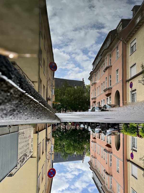 Street with yellow and orange-red three storey buildings, reflected in a puddle on the asphalt. The whole picture is turned upsidedown.