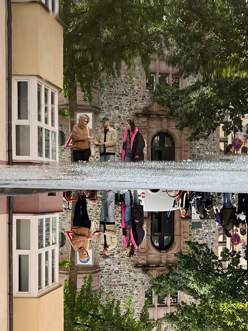 Street with yellow three storey buildings, on the left, and green trees. Three people in the middle of the street. The scene is reflected in a puddle on the asphalt. The whole picture is turned upsidedown. It looks like the three people Are standing in the water.