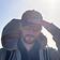 It's me, holding my cap in the wind. Wearing sunglasses. Short beard, slight smile. Sun in my back.