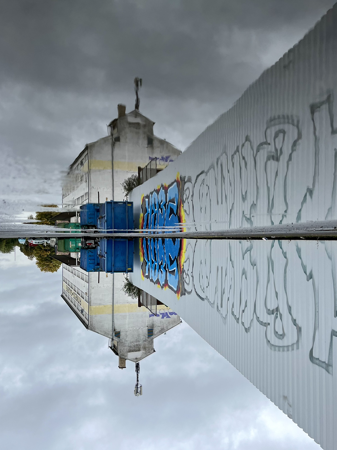 industry building, blue containers and a white wall with grafitti on it, all reflected in a puddle on the street. the whole picture is turned upsidedown.