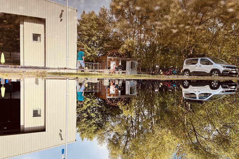 Flat white building with a big open Gate, Trees and a white car, all reflected in a puddle on asphalt. The whole picuture is turned upsidedown.