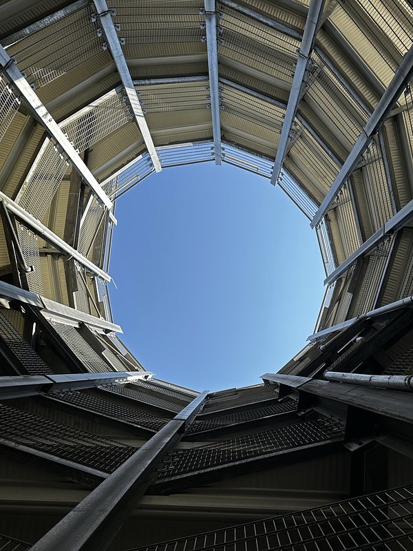 View from the bottom of a circular metal structure with mesh panels and support beams, looking up at a clear blue sky through an open top. The perspective creates a sense of depth and symmetry.