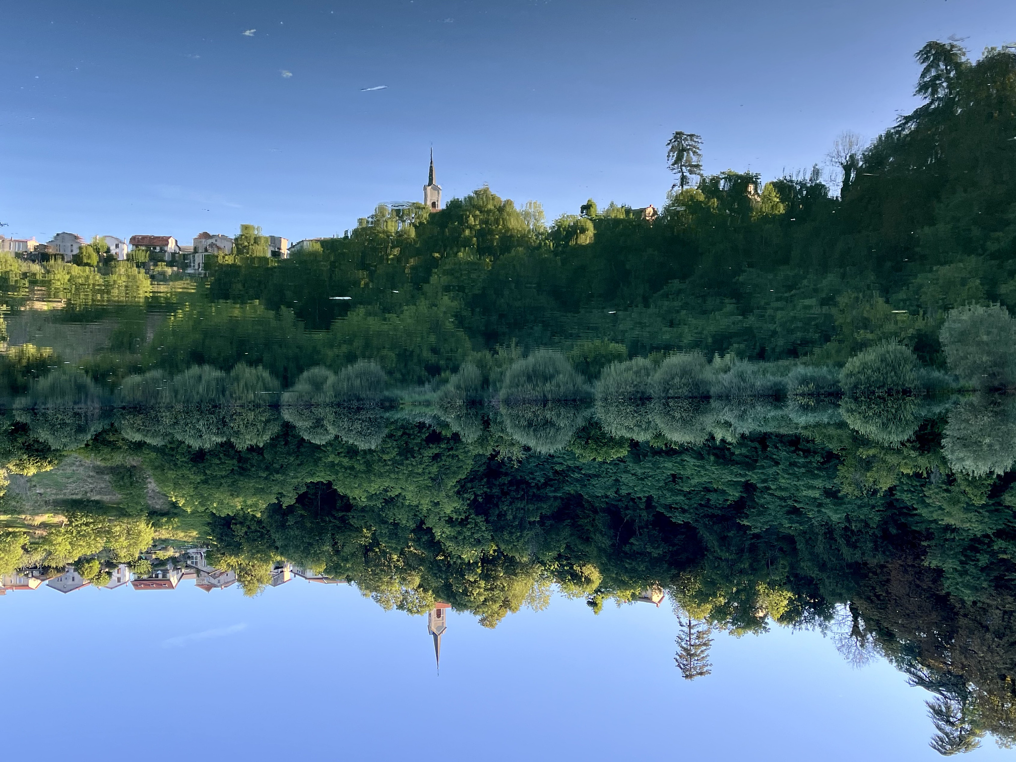 Green trees and bushes at a hill, reflected in a pond. At the top of the hill there is a church and some stone houses, also reflected in the pond. The picture is turned upsidedown.