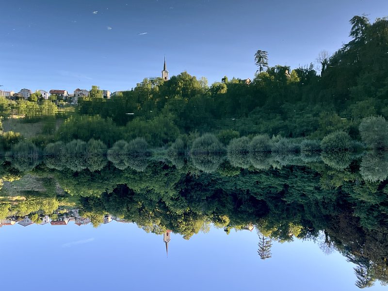 Green trees and bushes at a hill, reflected in a pond. At the top of the hill there is a church and some stone houses, also reflected in the pond. The picture is turned upsidedown.
