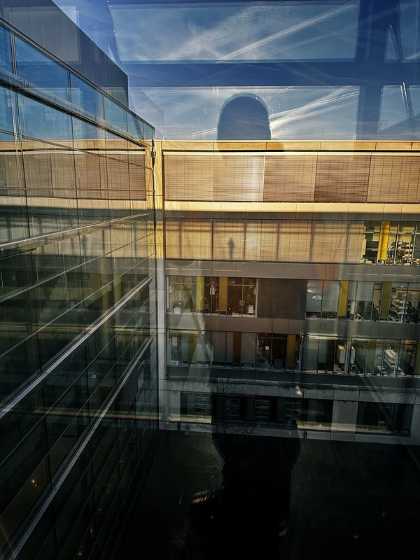 A reflective photo showing a modern glass office building with several floors. The windows reflect a blue sky with wispy clouds and contrails. The silhouette of a person is visible in the reflection, creating an abstract blend of interior and exterior elements. The interior office spaces have large windows with blinds, and some office furniture can be seen.