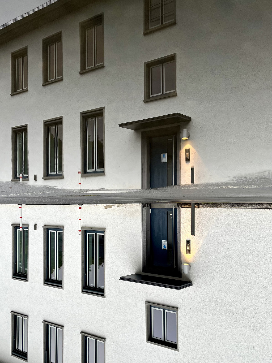 White building with blue door and a light next to the door, all reflected in a puddle. The picture is turned upsidedown.
