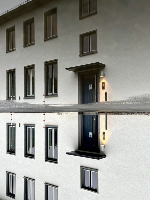 White building with blue door and a light next to the door, all reflected in a puddle. The picture is turned upsidedown.
