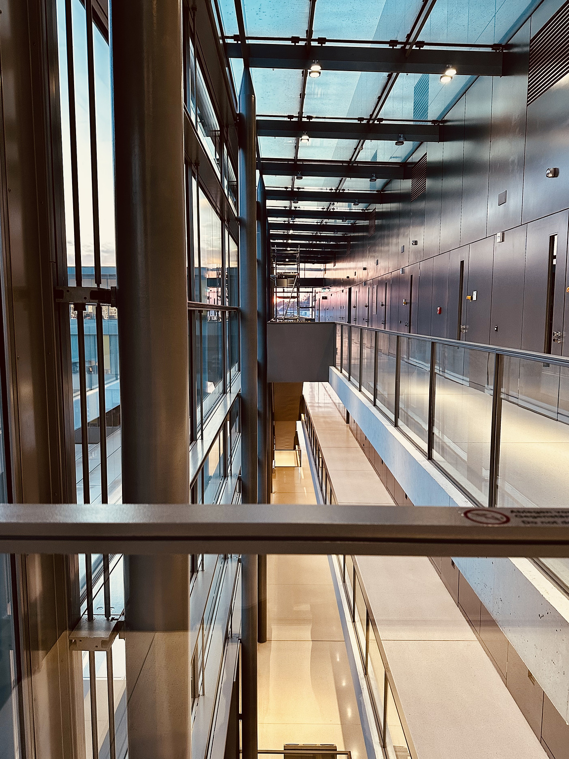 View of a modern, multi-level building interior with glass walls and a glass ceiling. The corridor features metal beams and railings, with reflections creating a sleek, industrial look. The lighting is soft, with a hint of the sunset visible through the windows. The design emphasizes open space and transparency.