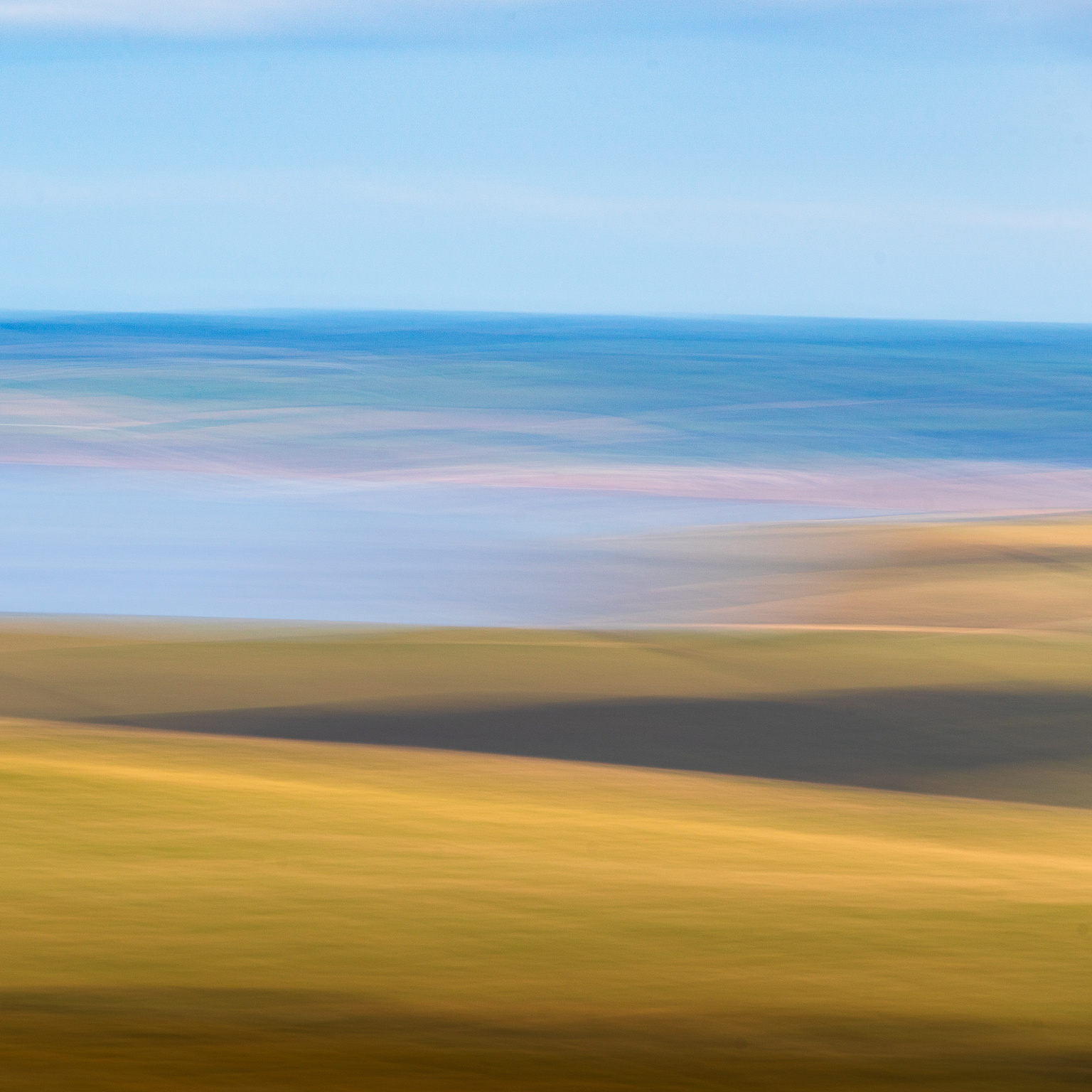 Square format abstracted landscape. Golden tones of autumn fields in the foreground at the bottom, in sweeping shapes describing the curves of the landscape. There is a band of pale blues of the sea, then above that the faded green and blues tones of the hills behind. Finally at the top there is a sky that is mainly blue but with suggestions of some white clouds.