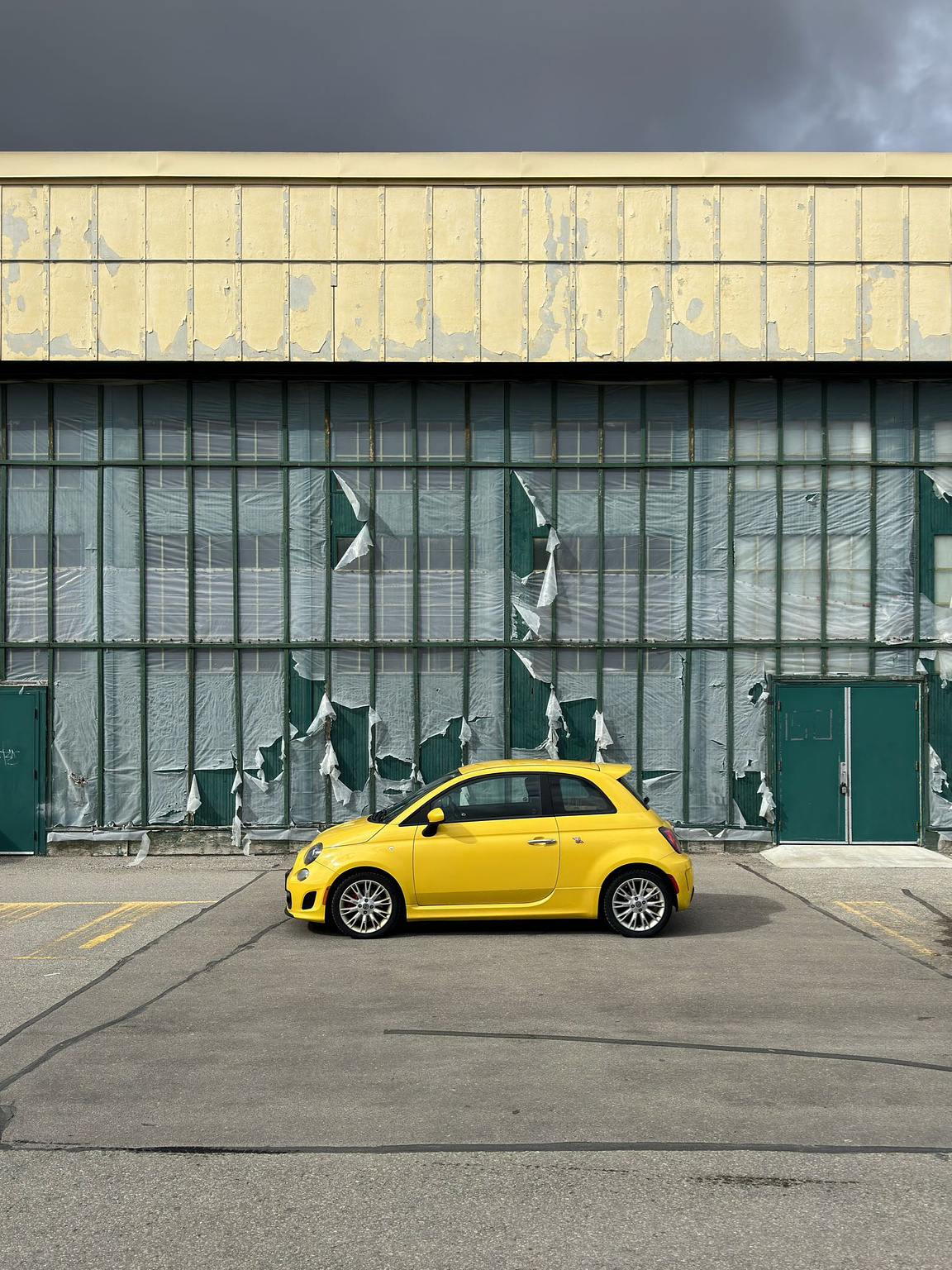 A tiny car, bright yellow, is parked in front of an abandoned warehouse. The deep green metal of its walls is covered in tattered white pastic sheets, fluttering in the wind. The top of the building is a wide sheet of faded yellow, mirroring the car below. Ominous clouds loom in the background.