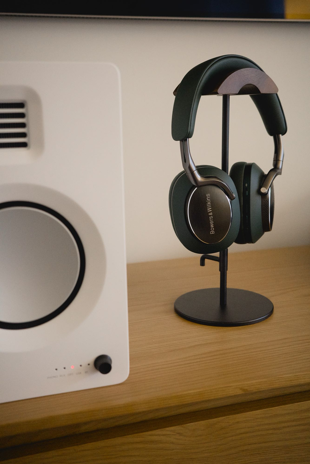 The Px8 on a headphone stand, seen beside a white bookshelf speaker on a media console.