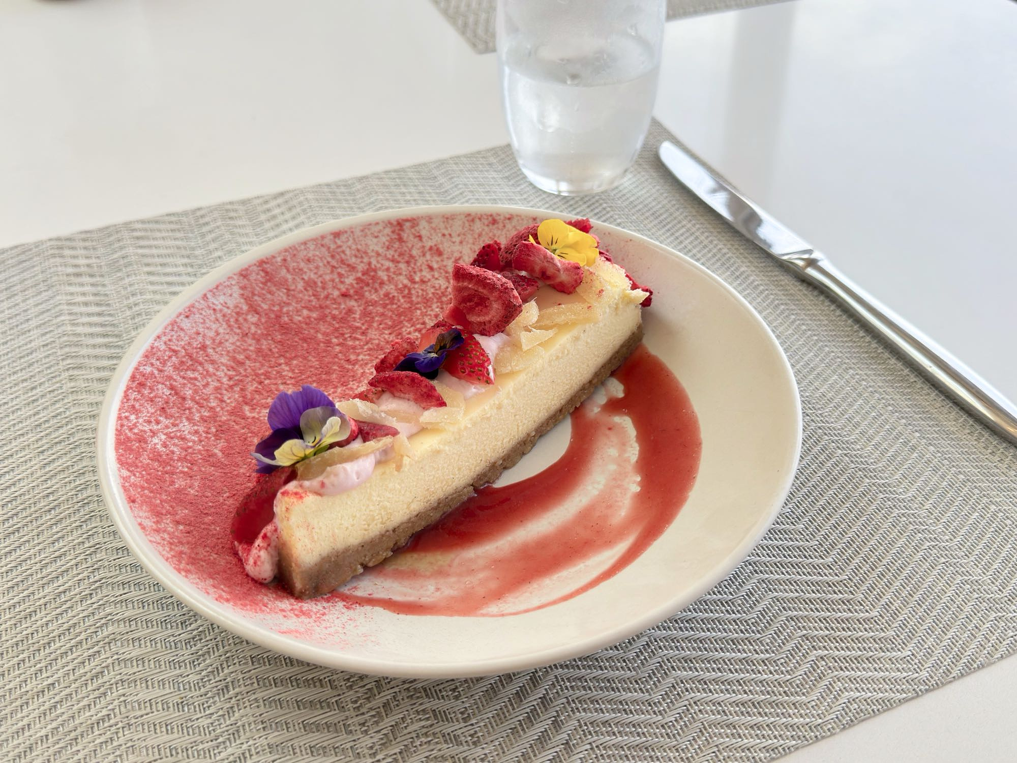 A fresh slice of New York style cheesecake sits on a small white plate, its surface swept with a gradient of decorative red powder on one side and a simple brushstroke of red jelly on the other. The cheesecake's surface is adorned with edible flowers and small pieces of fruit.