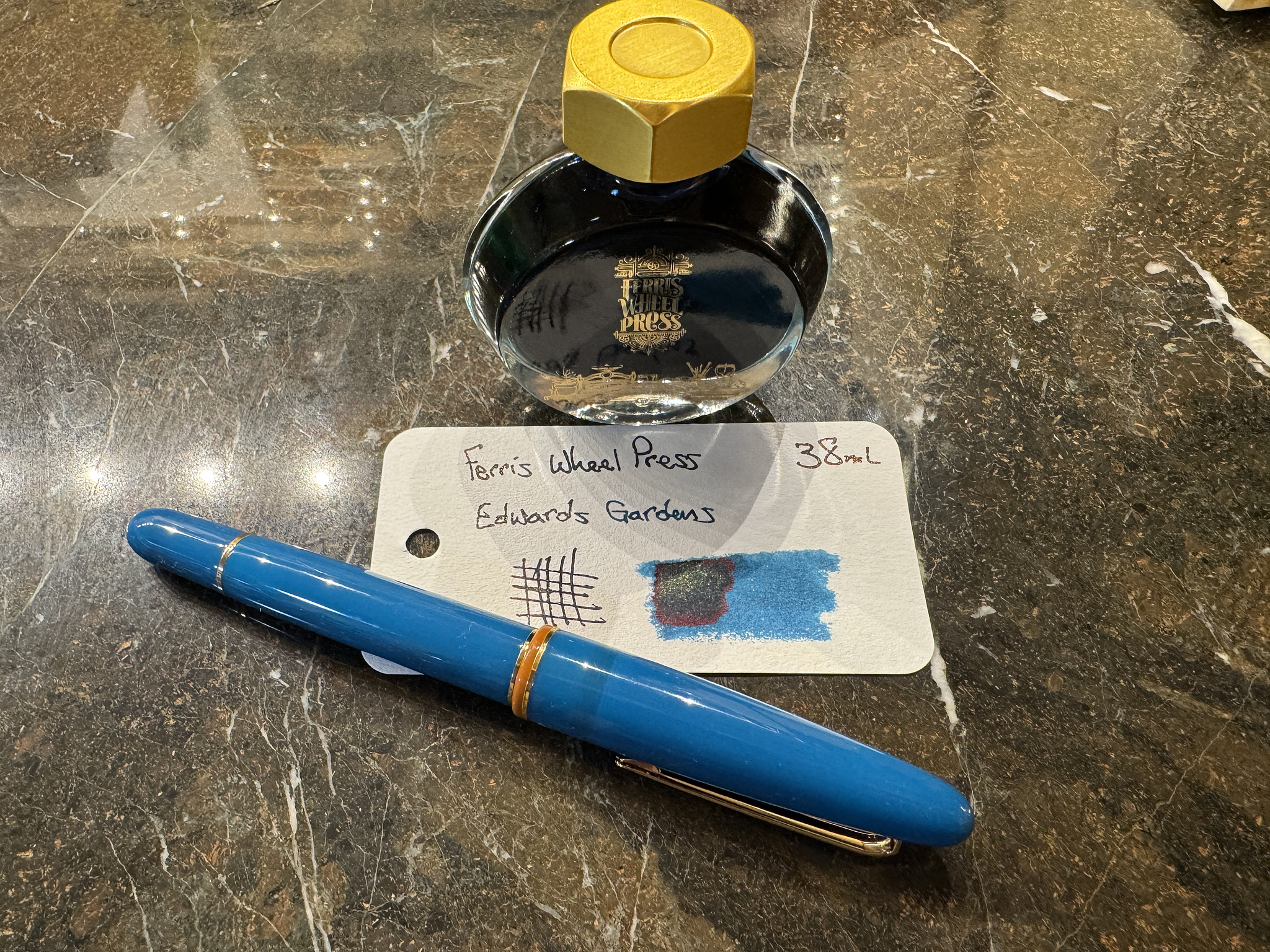 bottle of fountain pen ink with a gold cap and "Ferris Wheel Press" logo on the front, a swatch card showing a sample of the ink, and a blue Esterbrook Estie fountain pen capped; all sitting on a brown marble countertop