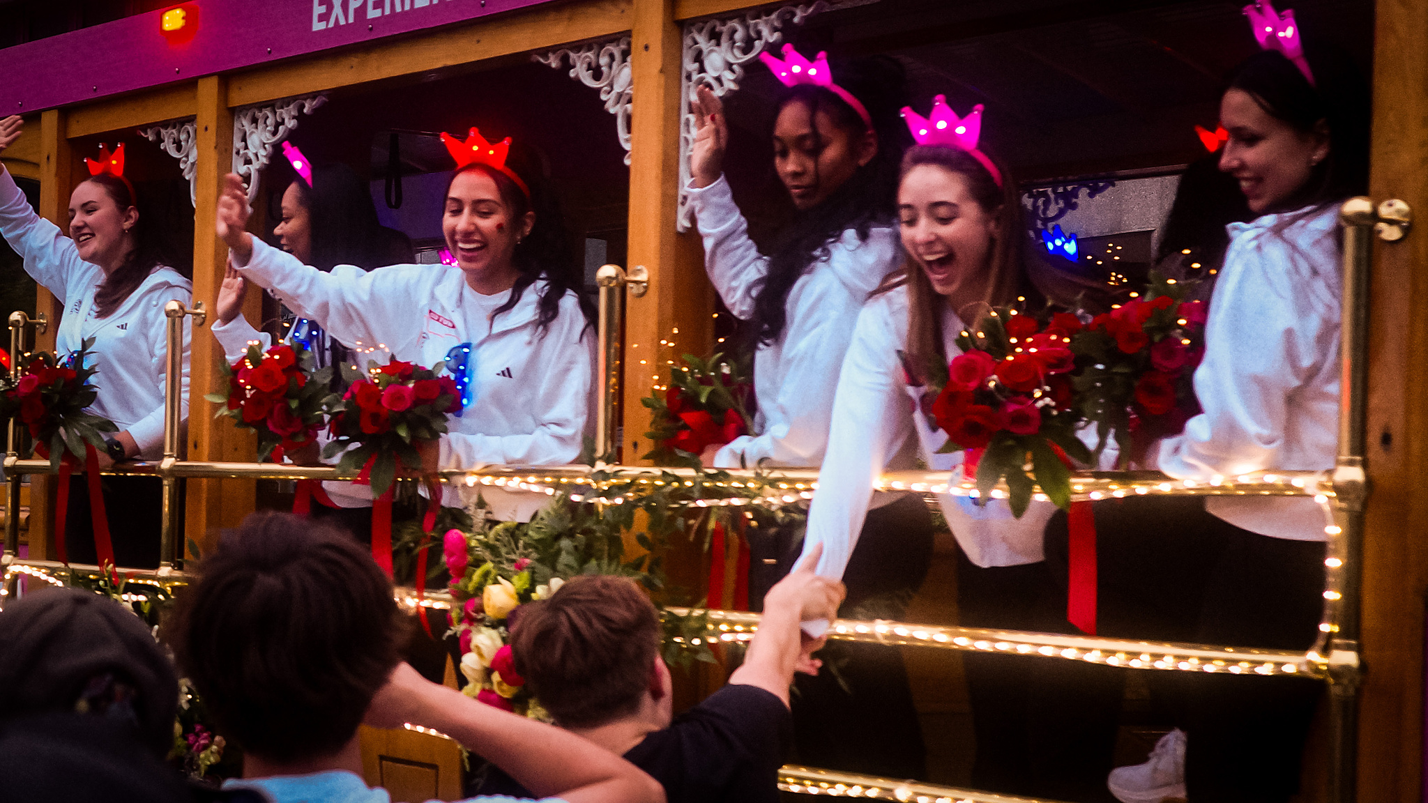 Young women in light-up tiaras wave and reach out to parade viewers from a streetcar. 