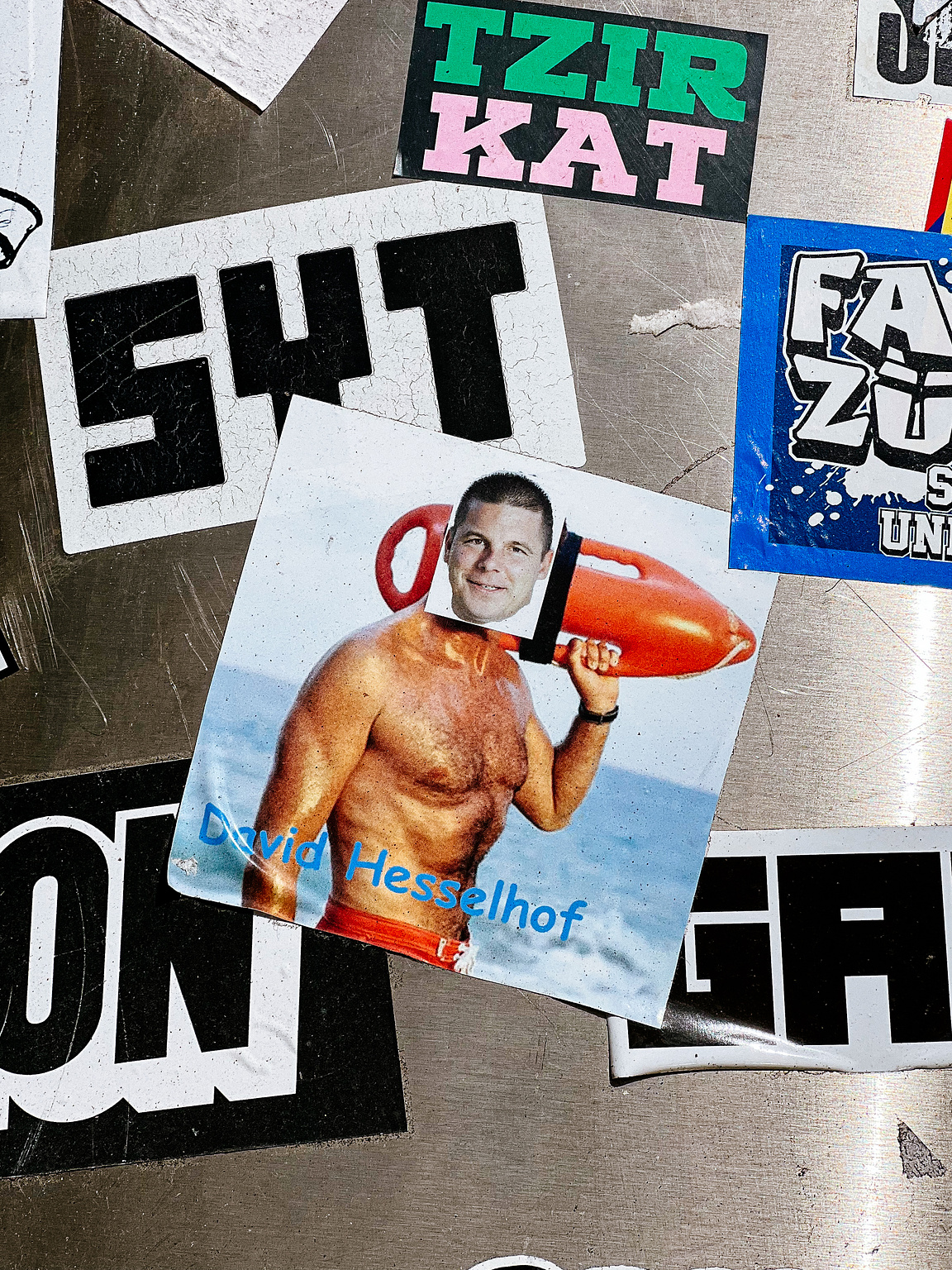 A collage of stickers on a surface, featuring a shirtless man holding a buoy, labeled "David Hesselhof," among other layered, partially obscured stickers.