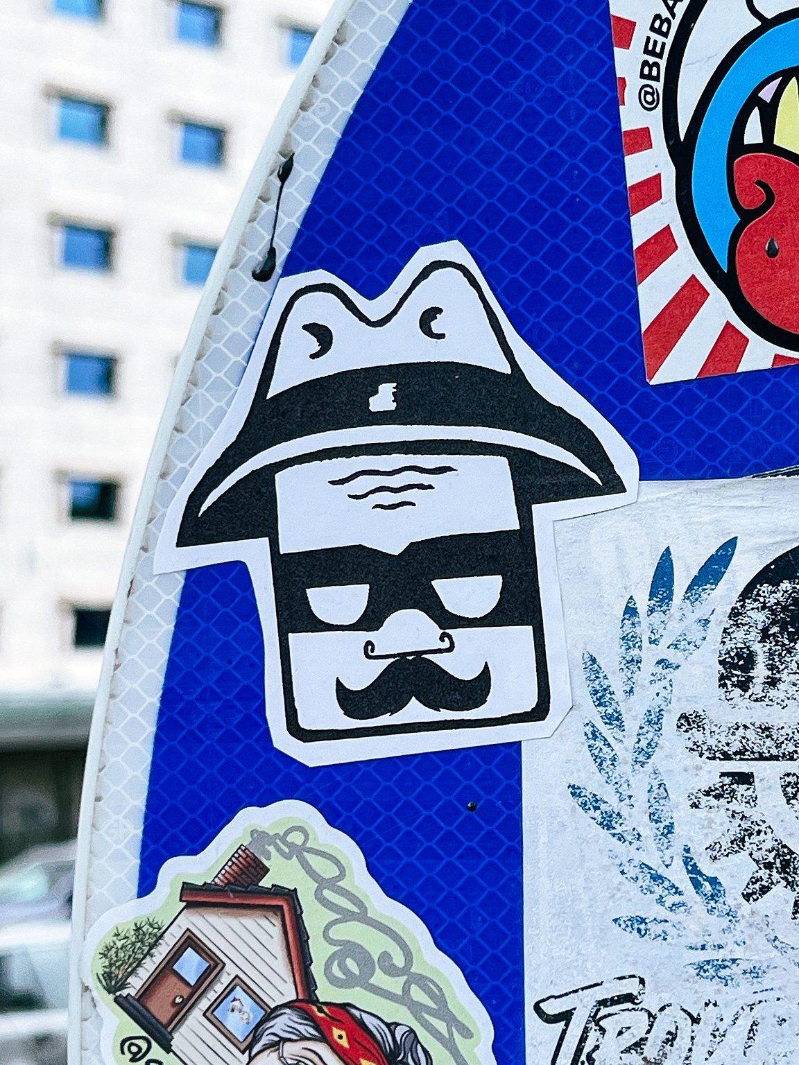 A sticker depicts a stylized face with a mustache and sunglasses, wearing a hat; it's adhered to a surface with other stickers, against a backdrop of buildings.