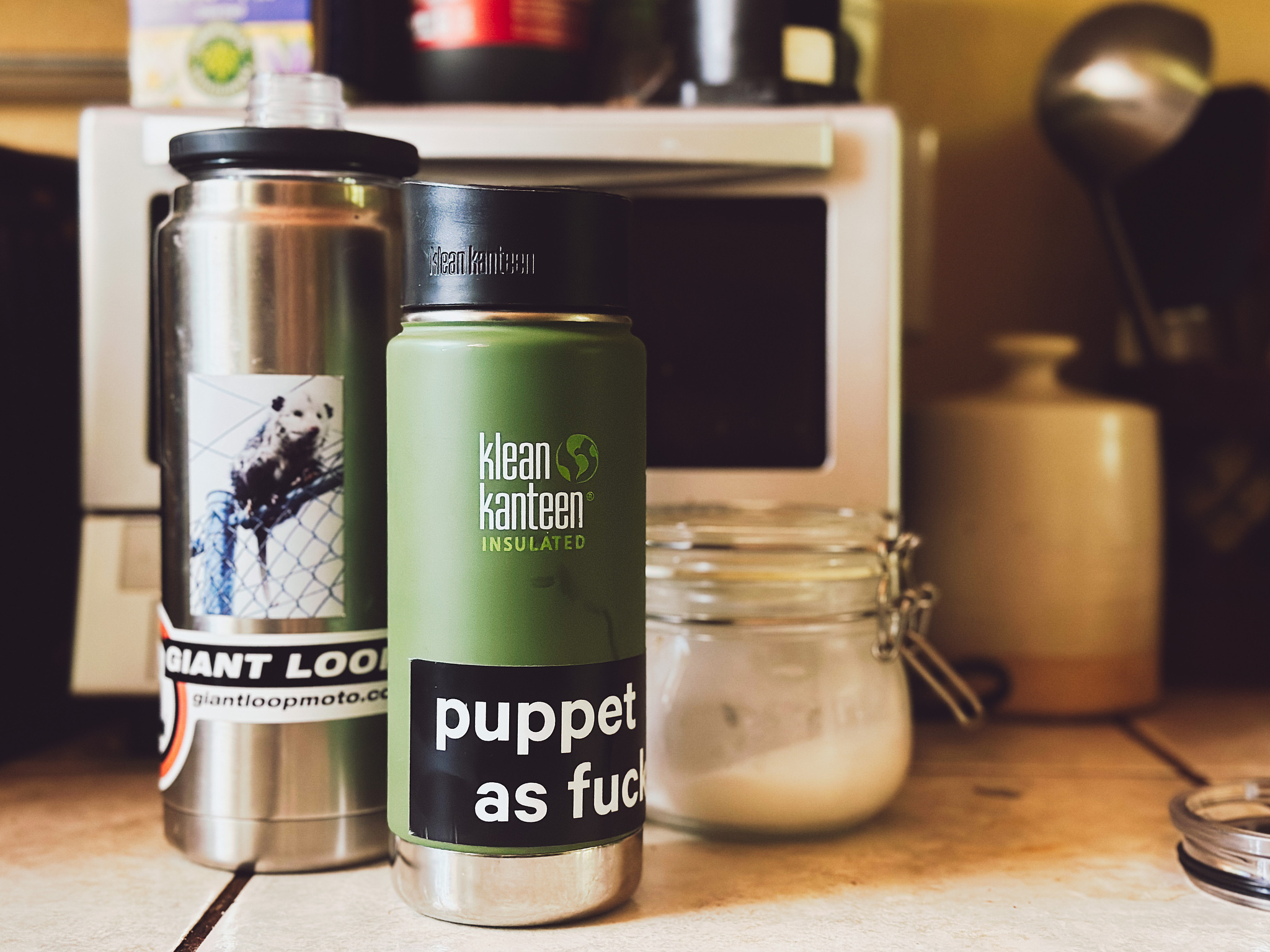 A green Kleen Kanteen with a sticker that reads "puppet as fuck." A silver water bottle with a photographic sticker of a possum.