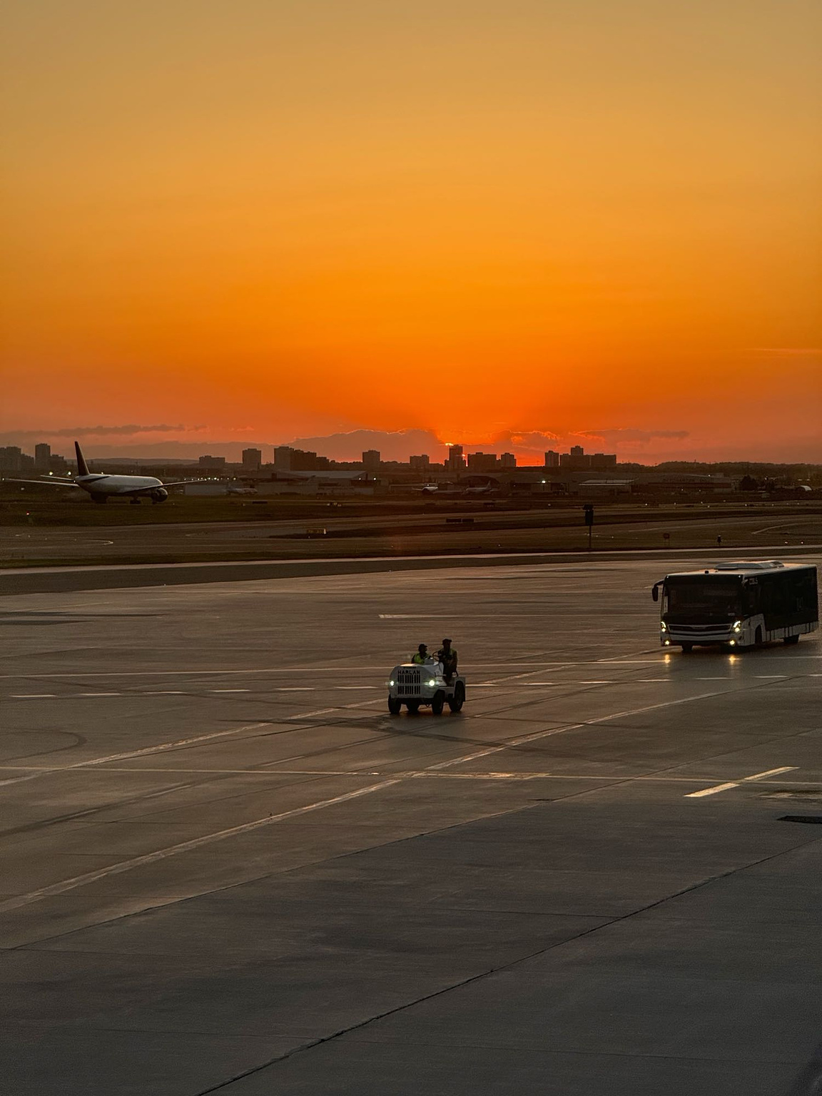 A telephoto view of an airport runway at sunset, with a baggage cart and passenger bus approaching in the foreground while a large commercial plane heads in the opposite direction in the background.