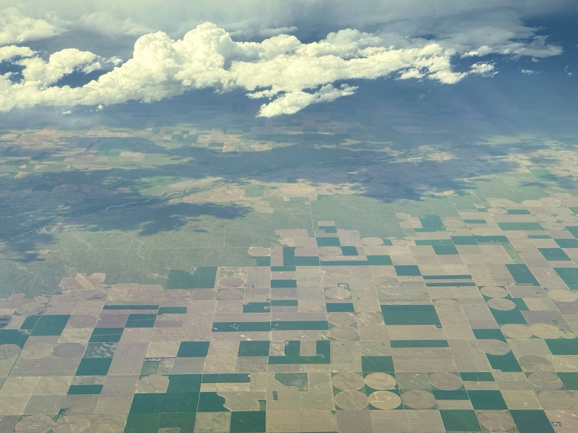 An aerial view of a rural landscape, dotted with square and circular parcels of land, each cultivating different crops. The result is a patchwork of green, yellow, brown, and gray over which a number of fluffy clouds float peacefully.