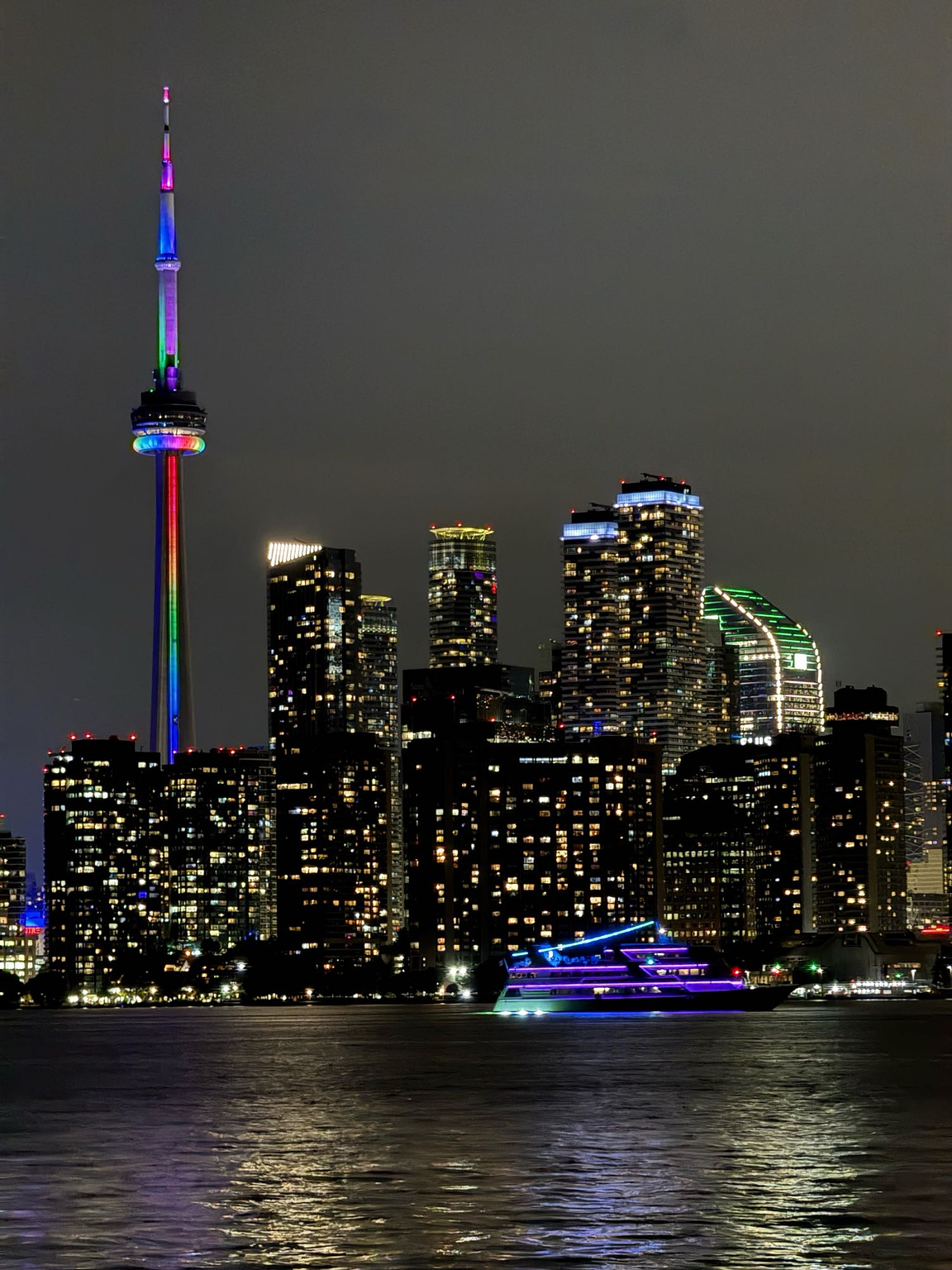 Neon lights on the CN Tower and its neighbouring skyscrapers in Downtown Toronto, seen from Toronto Island with a telephoto lens. A yacht cast in purple neon glow sails by in the foreground.