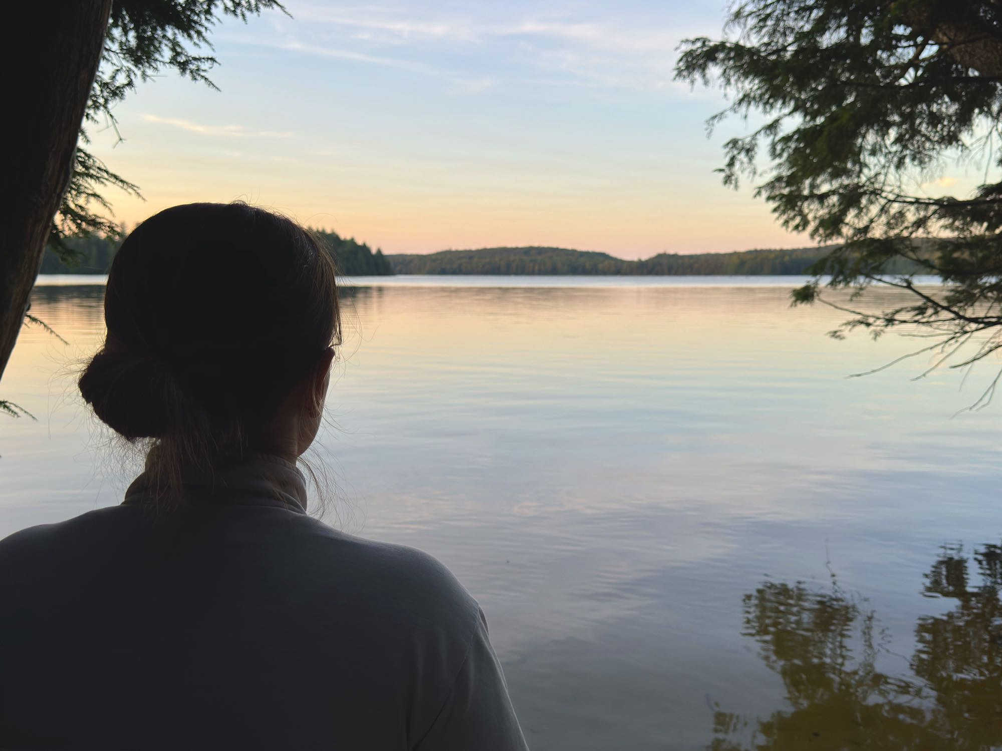 Sunset silhouette of a young woman standing in front of a peaceful lake, surrounded by deciduous trees. 