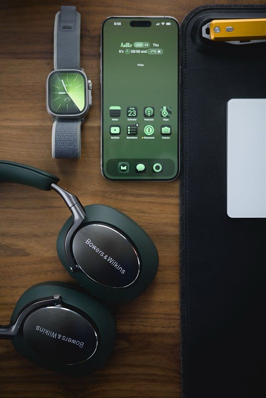 A flat-lay showcasing the Px8, an iPhone 15 Pro Max, and an Apple Watch Ultra 2. The phone and watch both have green wallpapers set up to match the colour of the headphones.