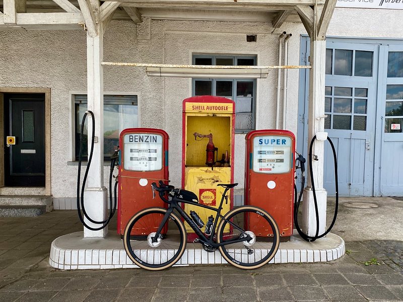 A gravel bike leans against vintage red Shell gas pumps in front of an old building.