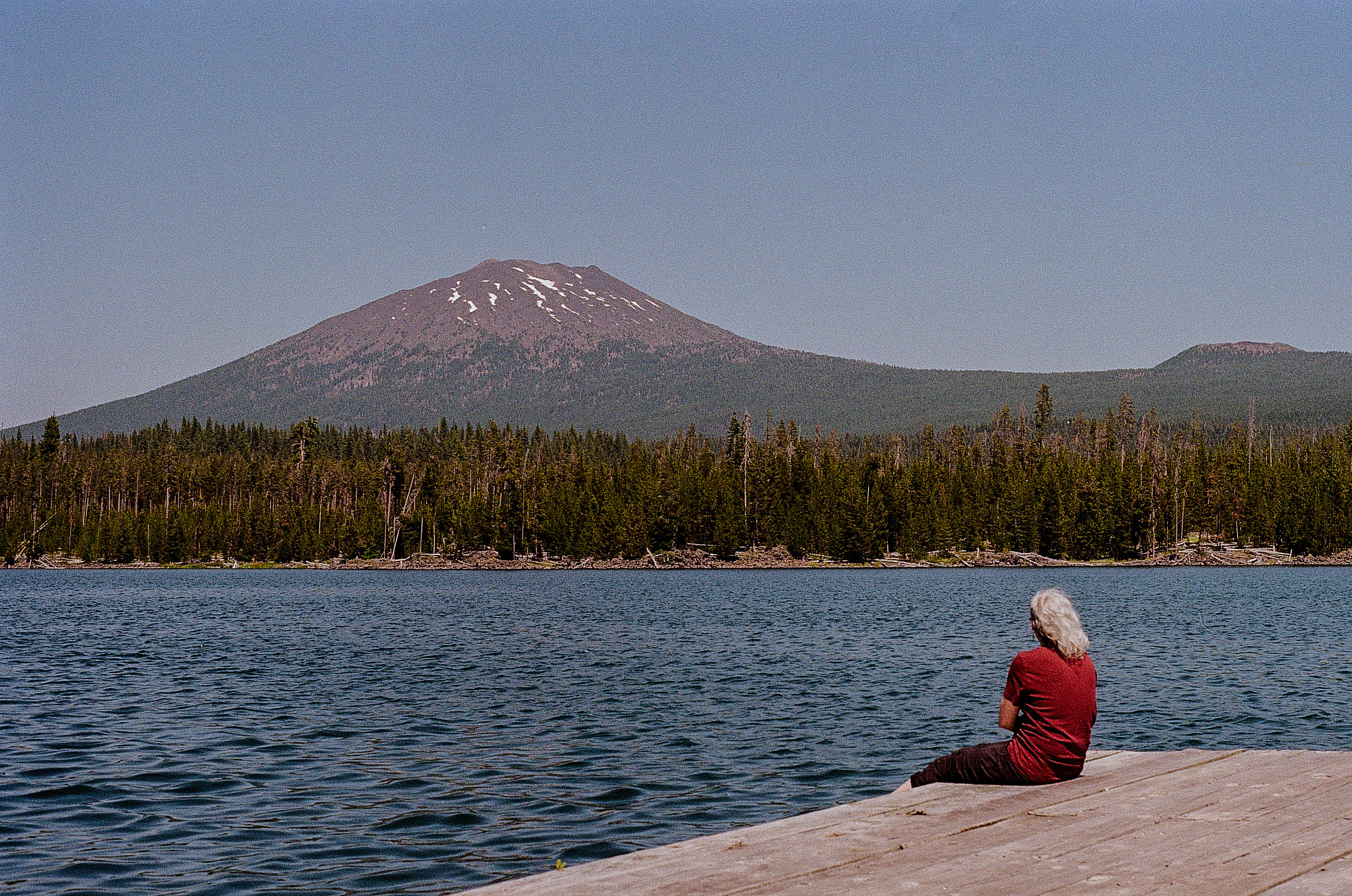 A woman with white hair in a dark red shirt sits on a dock on a mountain lake